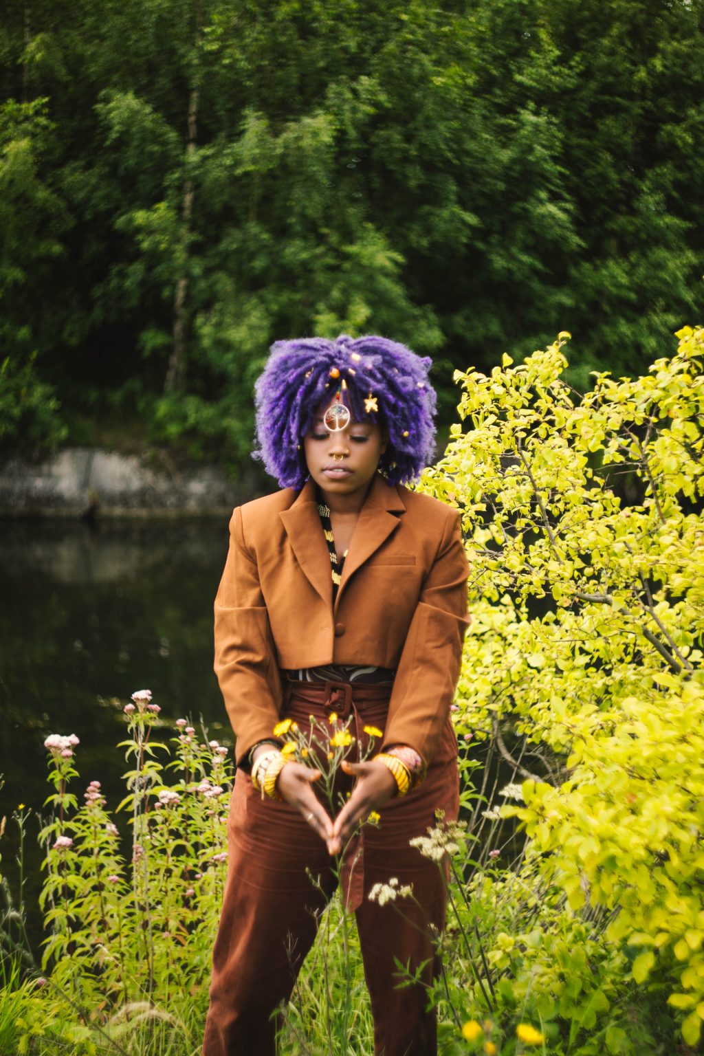 Eyve poses for a phot outdoors. There is a lake and tall trees in the background. Eyvehas a purple afro. She is wearing a brown suit and bangles. She has both hands in front of her with her fingers touching, forming an upside down triangle.