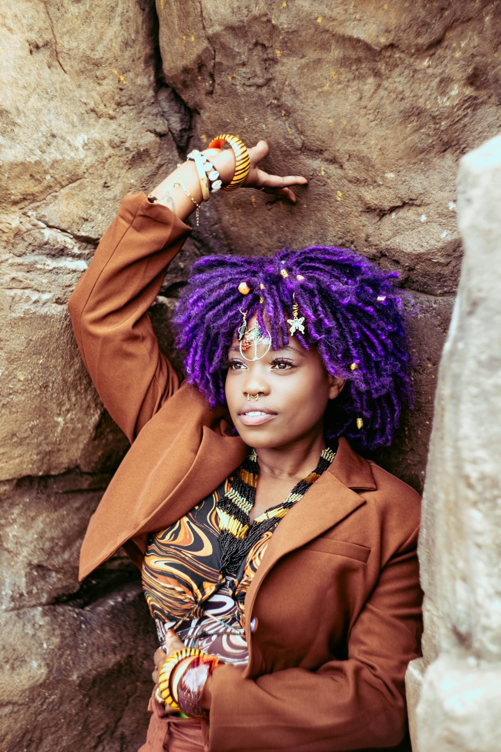 Eyve poses against a large wall of stone. She has a purple afro and is wearing a brown suit. She has one hand touching the tock above her head and another touching her torso.