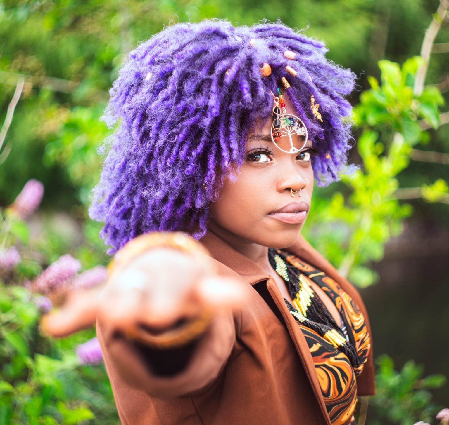 A close up portrait of Eyve. She has a purple afro and is wearing a charm which dangles over her face. Her hand is outstretched towards the camera.