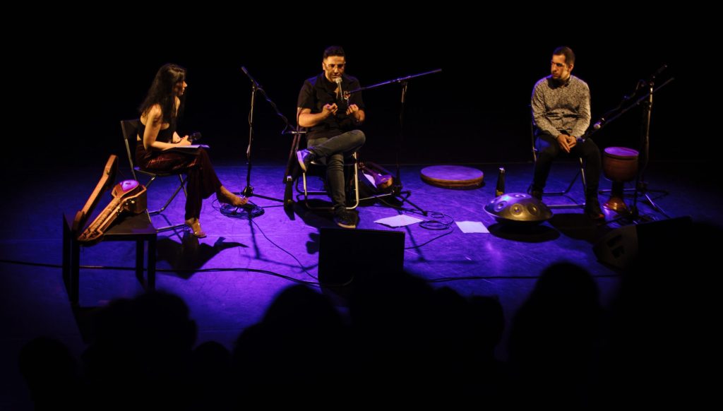 Three musicians are sat in a row on stage in front of an audience. They are illuminated by a stage light. Aref is sitting in the middle with his legs crossed and is speaking to the crowd using a microphone.