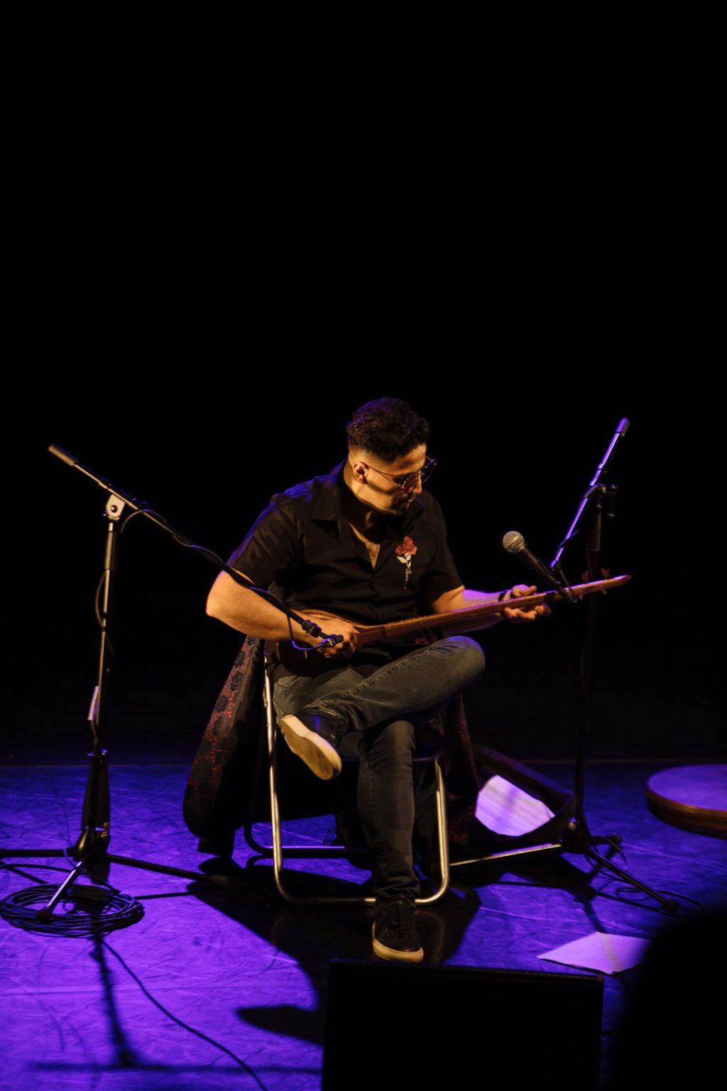 Edinburgh International Festival. Aref sits on a dark stage with his legs crossed as he plays the setar. There are two mics on stands in front of him.