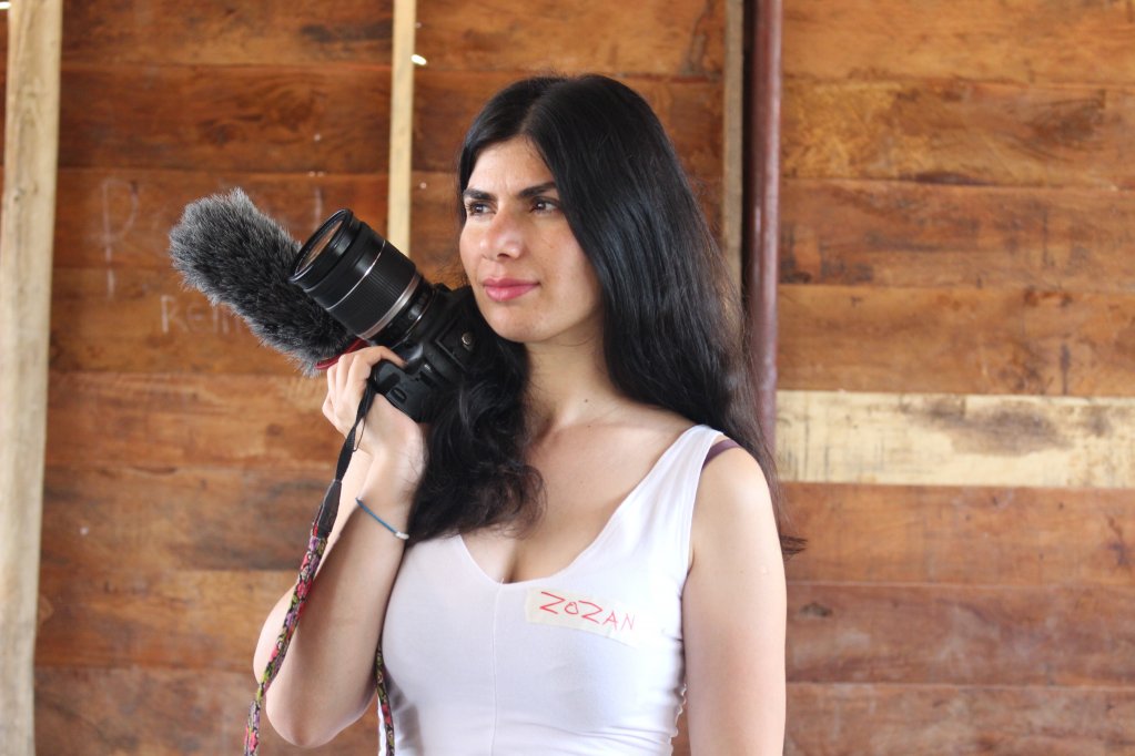 Zozan Yasar standing in front of a wooden wall. She has long dark hair. She is wearing a tank top and holding a large camera and microphone against her shoulder.