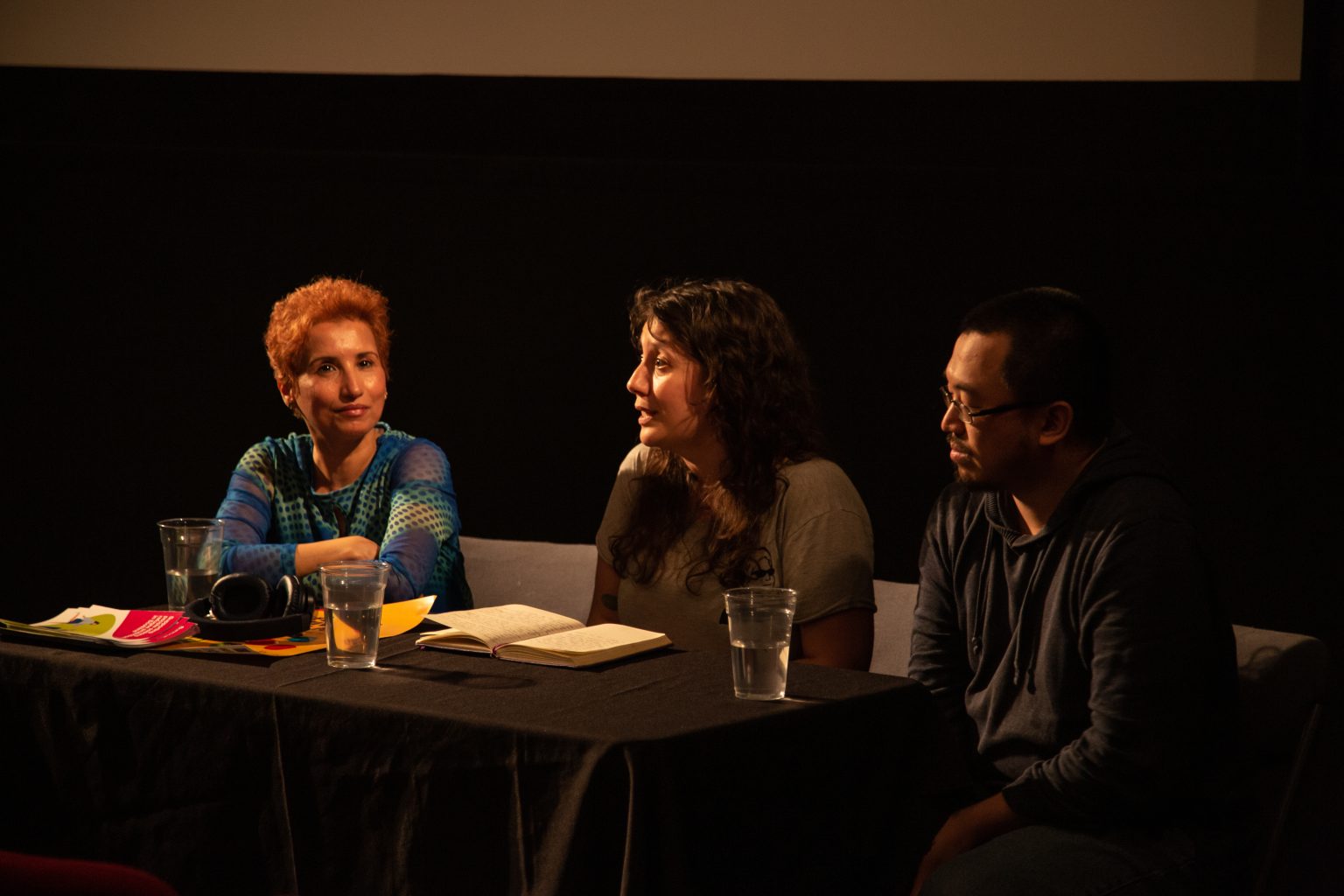 Three artists sat close together on a stage. They are taking part in a panel discussion. The artist in the centre is speaking to the crowd. The other two artists face her, smiling and listening. There are glasses of water, a headset and an open notebook on the table.