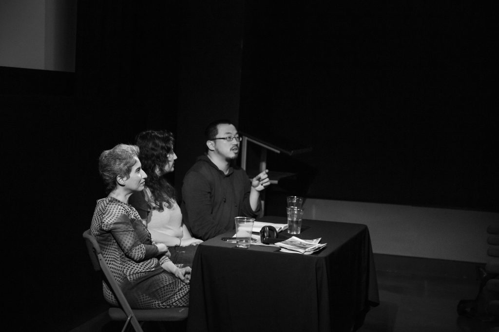 Three artists sat close together behind a small table covered by a table cloth. They are taking part in a panel discussion. The artist on the right is speaking to the crowd and gesturing with his hand. The other two artists face the crowd smiling and listening. There are glasses of water, a headset and an open notebook on the table.