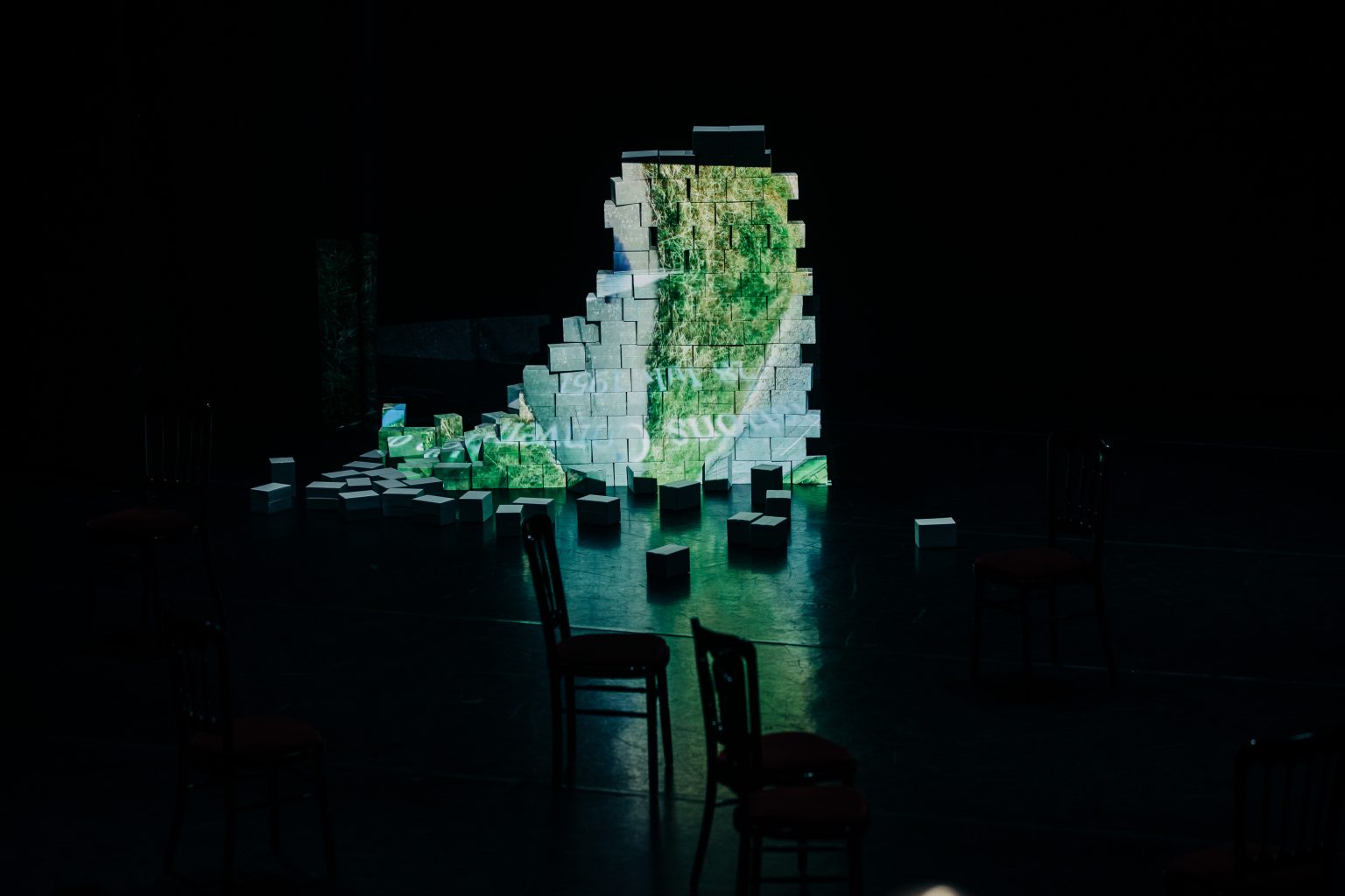 A half built and half demolished brick wall on a dark stage. There is an undiscernible image projected onto the wall.