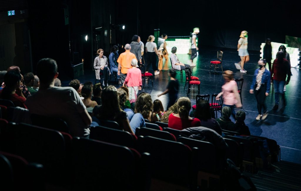 In the foreground, an audience sits in a dark theatre watching a staged production. On the stage, actors walk in a clockwise circle. There are several empty chairs scattered in the middle of the circle.