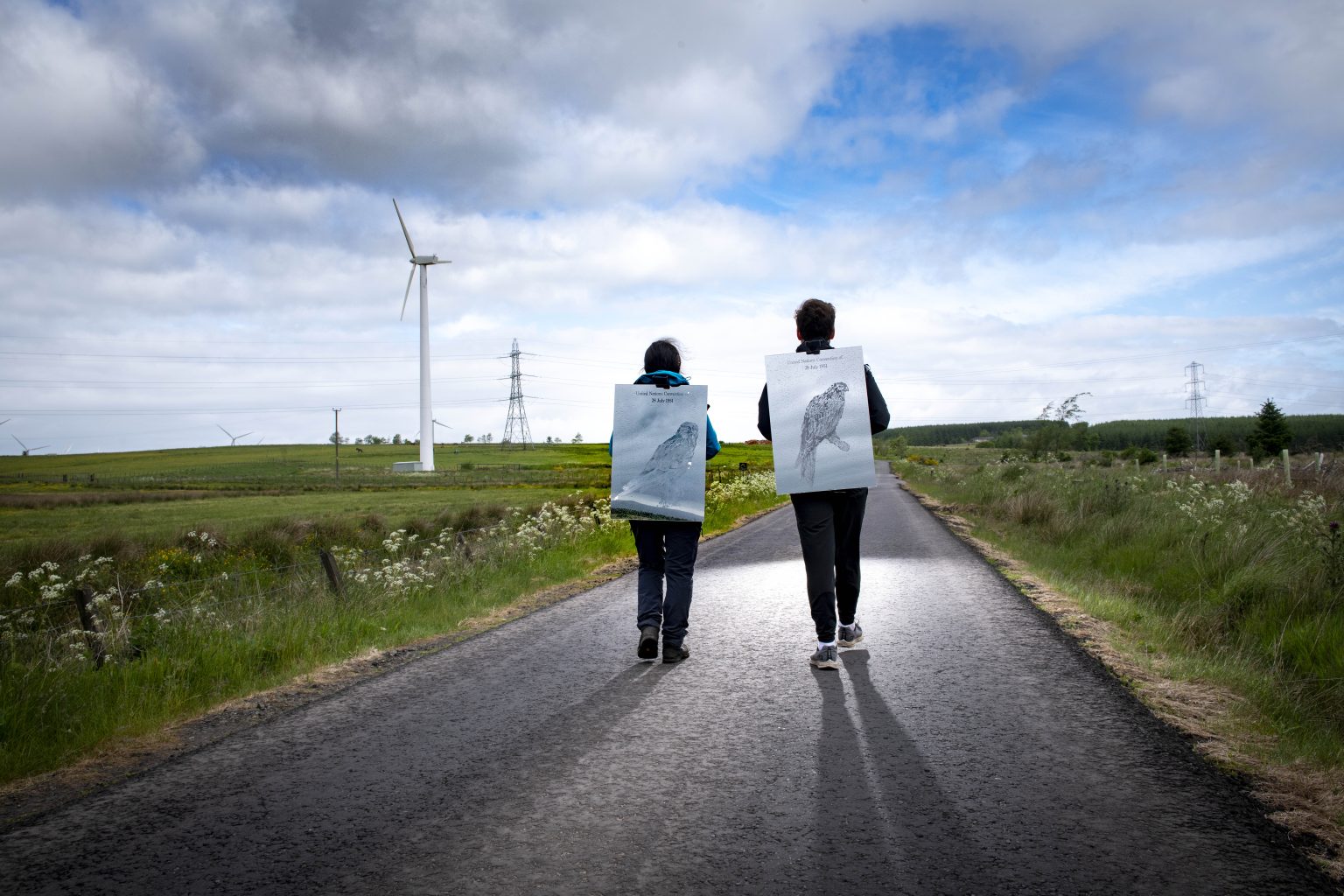 Two people walk down an empty country road wearing mirrors on their back. Each mirror has text and a carving of a bird. The road is surrounded by fields of grass. There are windmills and phone towers in the background.