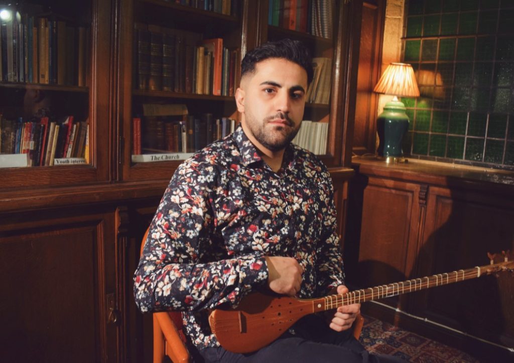 Aref Ghorbani posing in front of a wooden bookcase. He has dark hair and facial hair, is wearing a floral shirt and is sitting on a wooden chair. He is holding a setar, a small and long string instrument made from wood.