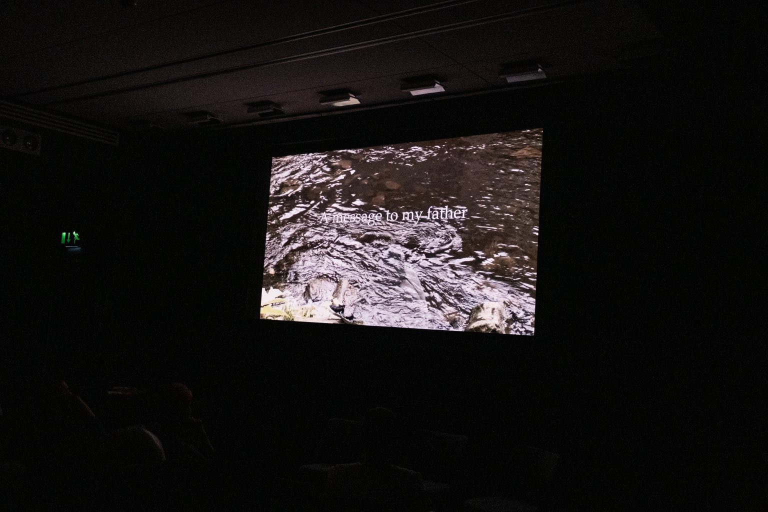A cinema screen showing a scene from a film. On the screen there is an image of a body of flowing water and text which says 'A message to my father'.