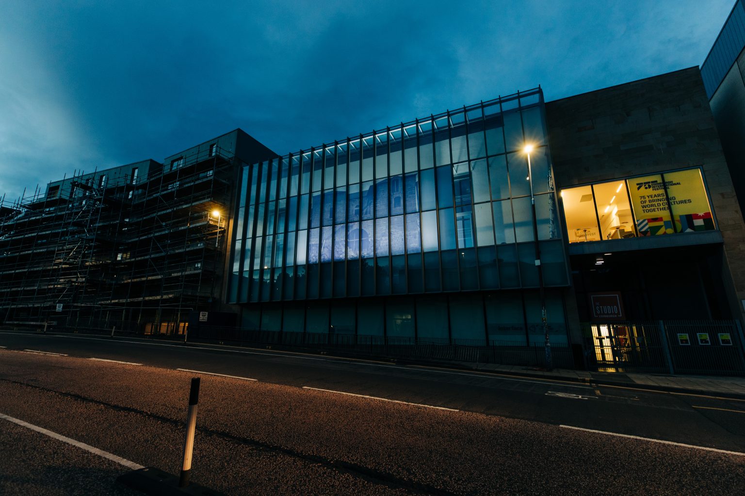 A modern building in Edinburgh at dusk. The facade is made of glass and iron. On the left of the building there is a poster advertising Edinburgh International Festival.