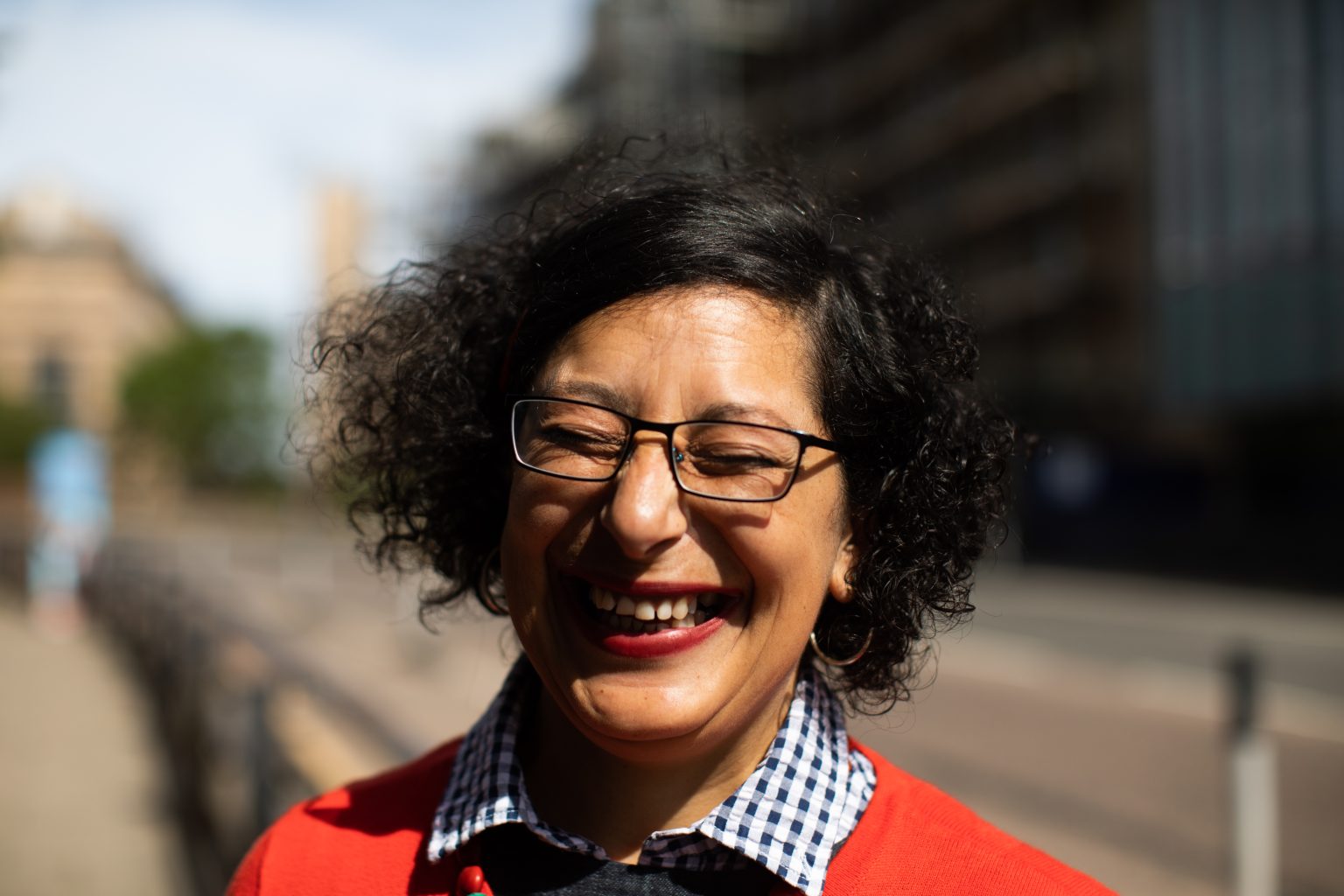 A close up of Leena Nammari standing on a city street. She has short curly hair and is wearing glasses and a collared shirt. She is facing the camera and smiling with her eyes closed.