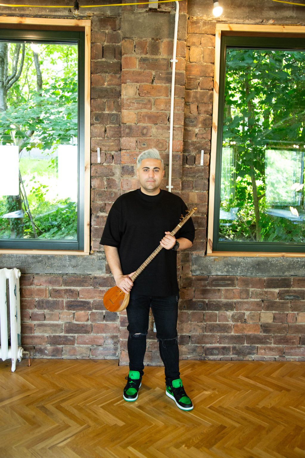 Aref stands in front of a brick wall. He is wearing a t-shirt and jeans and colourful runners. He faces the camera holding a string instrument in both hands.