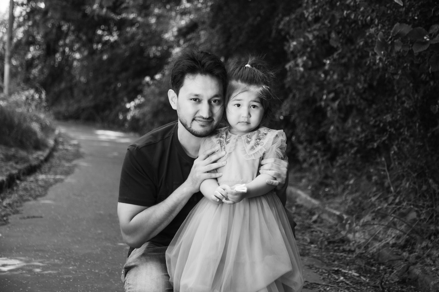A man is crouched beside his young daughter on a narrow road. They are both smiling towards the camera. His daughter wears a tulle dress while he wears a t-shirt and jeans.