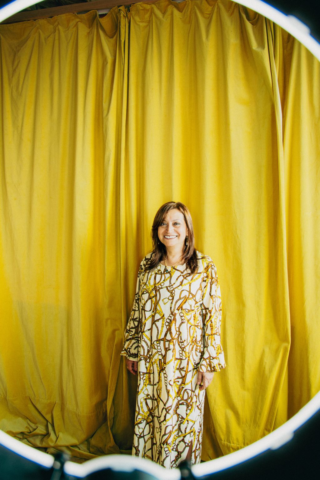 A woman stands in front of a yellow curtain. She is wearing gold jewellery and is smiling broadly. There is a white ring around the frame.