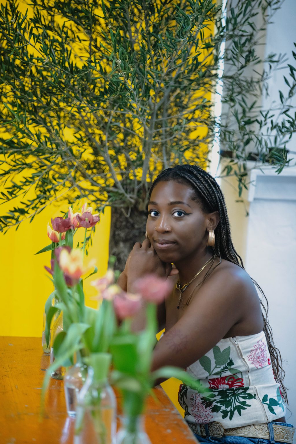 A black woman with braided hair poses on an indoor picnic bench. There is a large houseplant behind her and several glasses vases of tulips in front of her. She is leaning on the table top with her head turned over her shoulder and towards the camera.