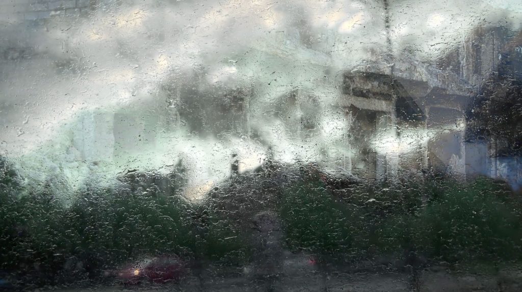 A blurred photograph of a rain drenched window. An indiscernible building is visible in the reflection.